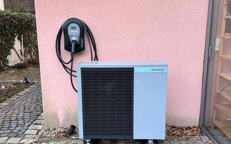 Wärmepumpe mit angeschlossener Wallbox vor rosafarbener Hauswand auf gepflastertem Hof
